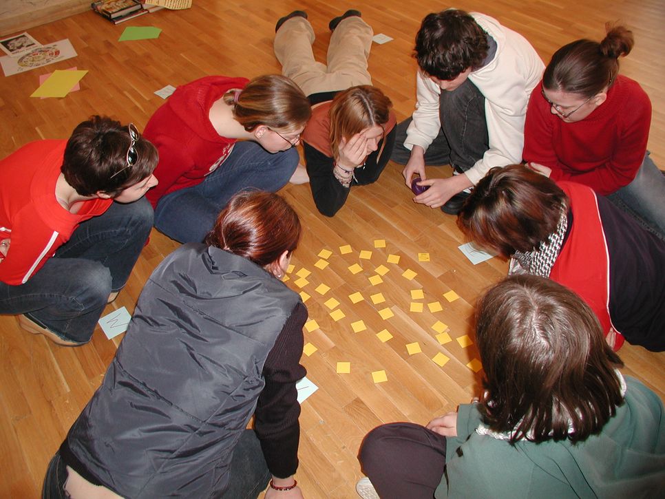 Jugendliche GruppenleiterInnen sitzen und teilweise liegen im Kreis. In der Mitte liegen kleine orange quadratische Zettelchen.