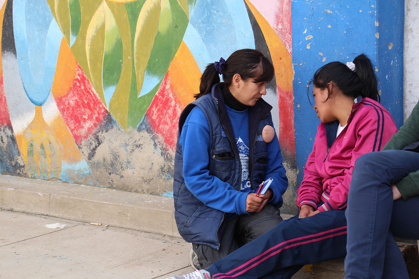 Bolivien: Alternativen zur Straße/Bild: DKA Eine Frau sitzt am Boden an eine Mauer gelehnt. Eine andere Frau sitzt kniend neben ihr und spricht mit ihr oder hört ihr zu.