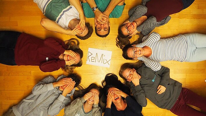 Das Regionalteam liegt am Rücken im Kreis und lacht. Jeder hat seine Hände entweder auf seine Augen, Ohren oder seinen Mund gelegt. In der Mitte des Kreises liegt ein Zettel auf dem ReMix steht.