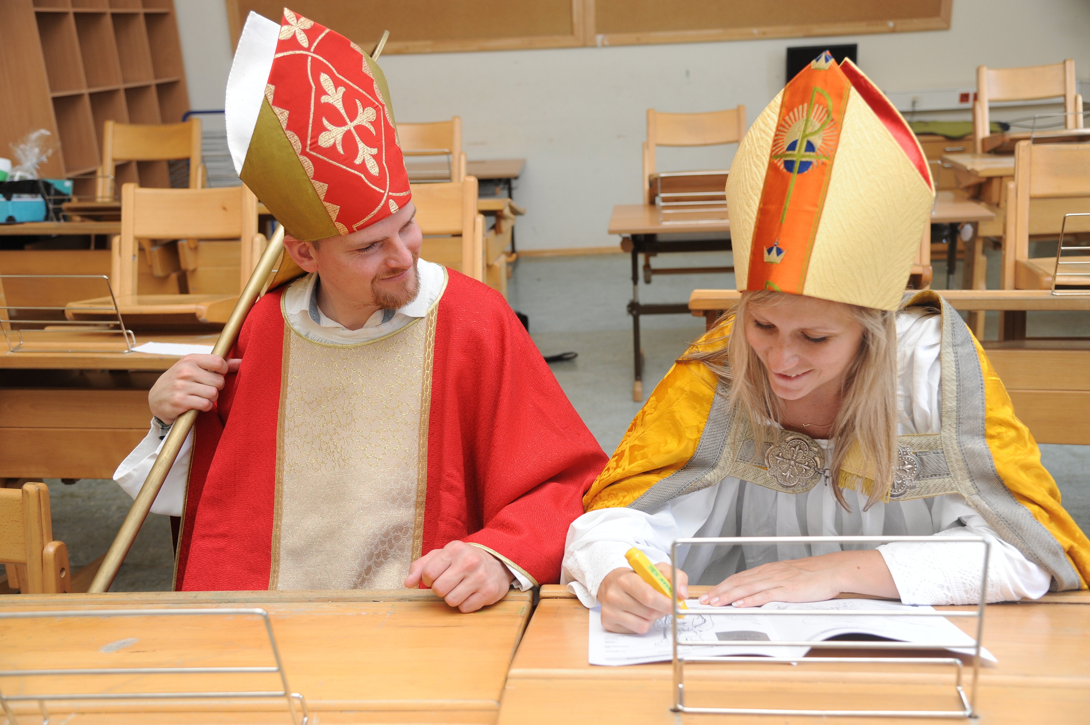 Credit: Kath. Jungschar St. Pölten Zwei verkleidete Nikoläuse sitzen nebeneinander auf einer Schulbank in einem leeren Klassenzimmer