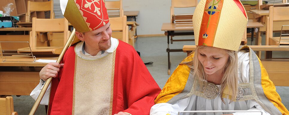 Zwei verkleidete Nikoläuse sitzen nebeneinander auf einer Schulbank in einem leeren Klassenzimmer