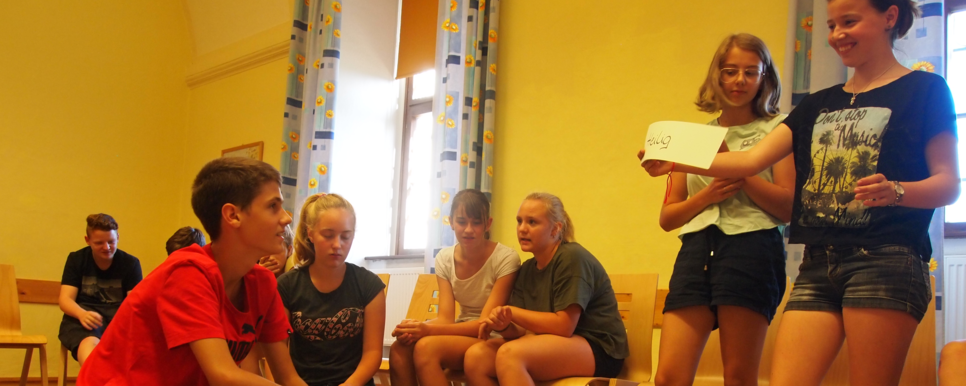 Credit: Kath. Jungschar St. Pölten Das Bild zeigt eine Schulung für Jugendliche. Einige Jugendliche sitzen auf Sesseln im Kreis. Andere hocken bzw. stehen in der Mitte auf einem Teppich und besprechen Kärtchen auf denen einzelne Begriffe stehen.