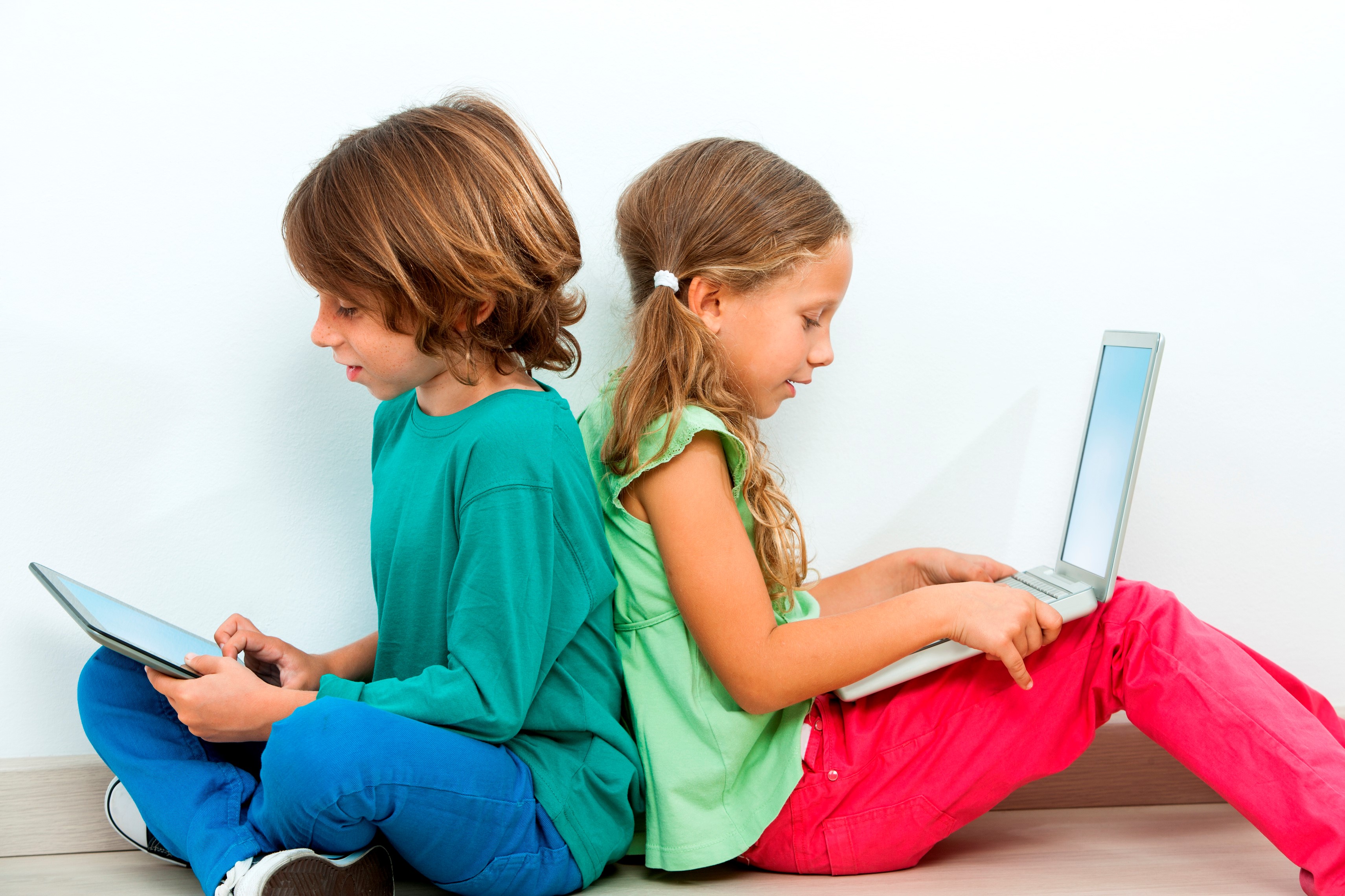 Zwei Kinder sitzen, Rücken an Rücken, auf dem Boden. Das Mädchen hat einen Laptop am Schoß und der Junge hält ein Tablet in der Hand.