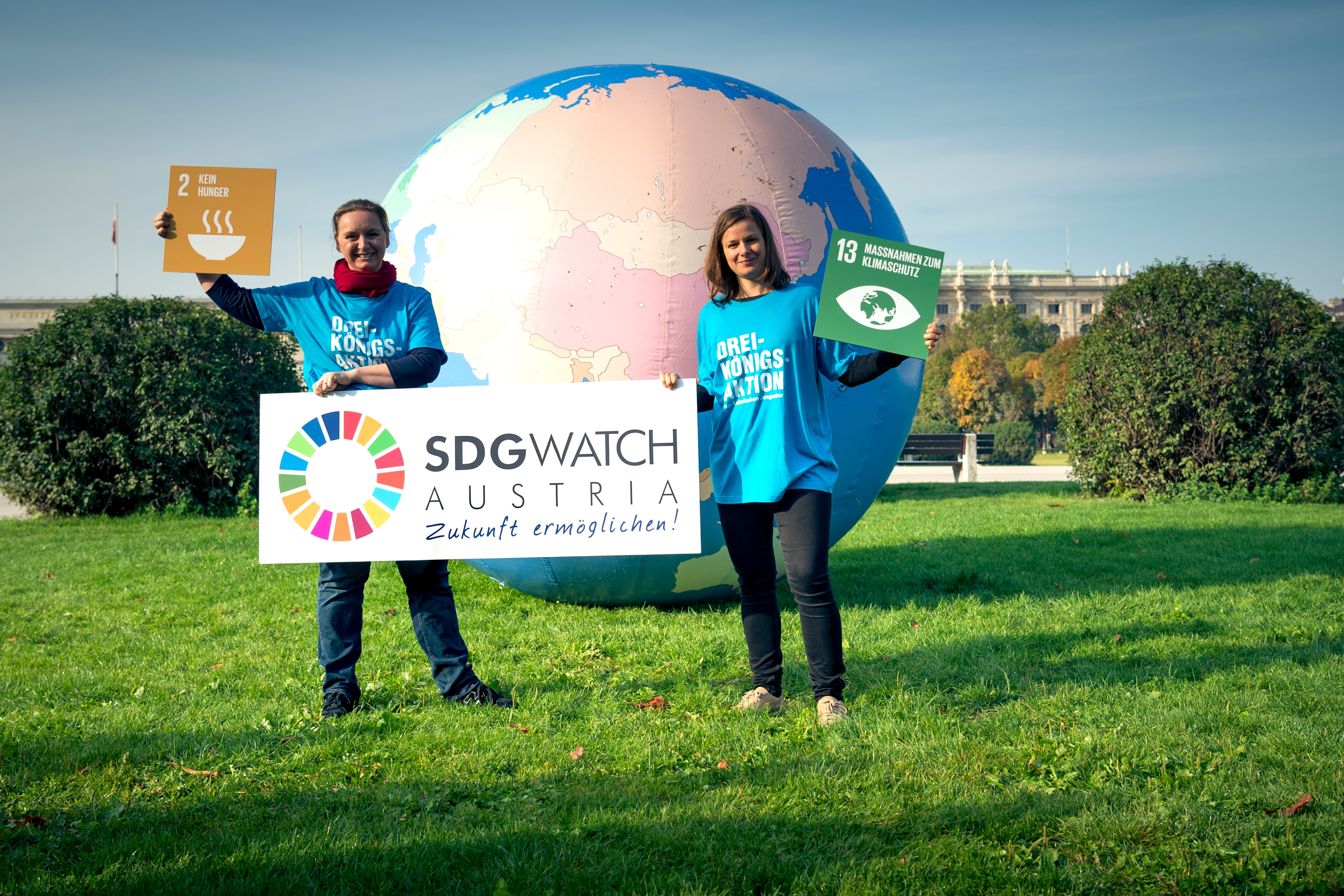 Zwei Angestellte aus der Dreikönigsaktion halten ein Schild "SDG Watch Austria" und Schilder der SDG Ziele "2 Kein Hunger" und "13 Maßnahmen zum Klimaschutz". Hinter den beiden ist ein riesiger aufgeblasener Globus.