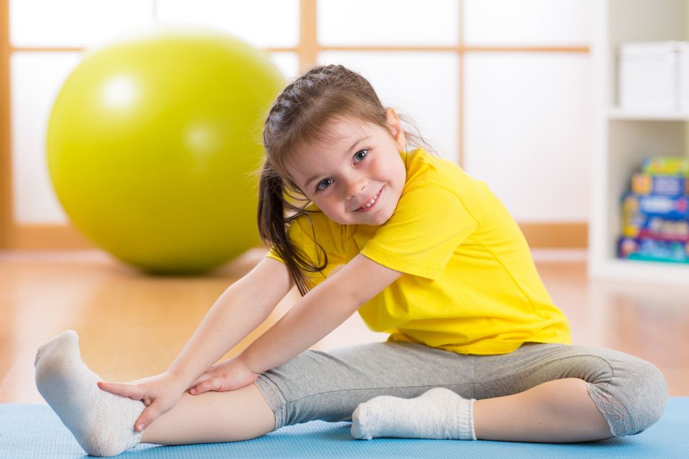 Kind im gelben T-Shirt sitzt auf Yogamatte und macht Turnübungen. Dahinter ist ein gelber Gymnastikball.