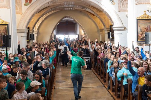 Kinder sitzen in den Bänken und halten beide Arme in die Höhe. Im Mittelgang gehen die Mitarbeiter*innen durch und animieren die Kinder.