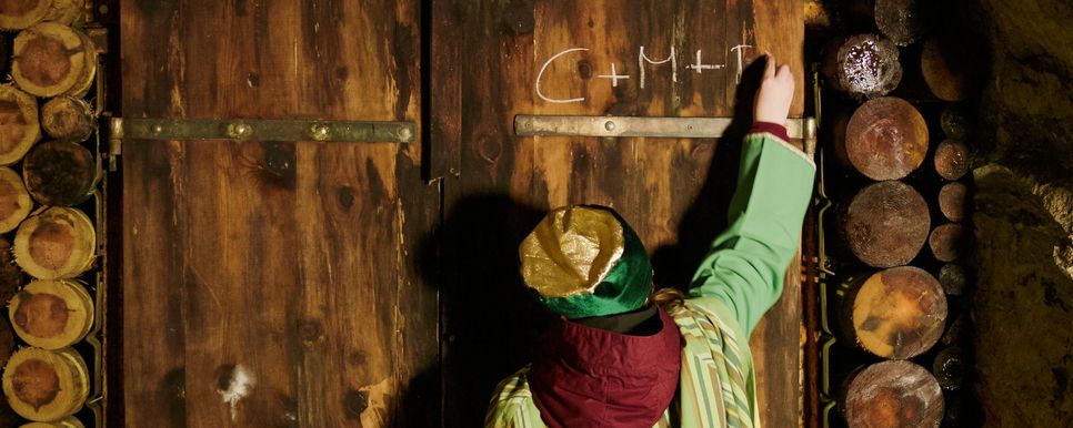Ein Sternsingerkind schreibt den Sternsingersegen mit Kreide auf eine Holztür.