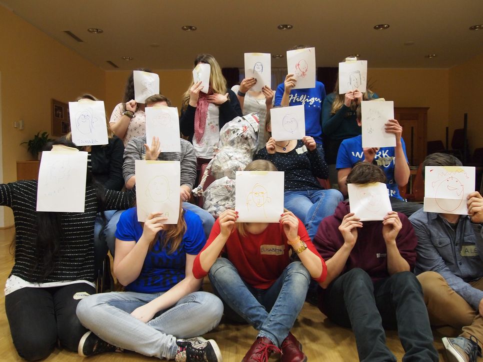 Jugendliche sitzen in drei Reihen hintereinander. Jeder/Jede hält ein Blatt Papier vor das Gesicht. Auf diesem Papier ist wiederum ein Gesicht gemalt.