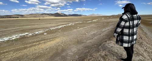 Eine indigene Frau steht am Rand einer flachen ausgetrockneten Ebene.