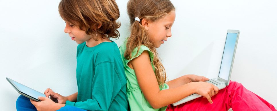 Zwei Kinder sitzen mit dem Rücken gegeneinander auf dem Boden. Das Mädchen hat einen Laptop auf dem Schoß. Der Junge hält ein Tablet.