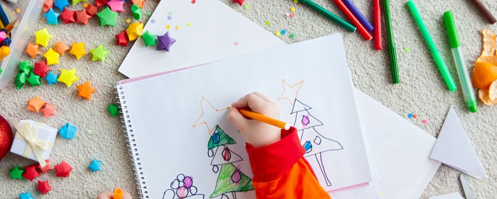 Zwei Kinderhände halten Stifte in der Hand und machen Bäume auf einen Block. Rundherum liegen Stifte, Bastelmaterialien und kleine Sterne aus Papier.